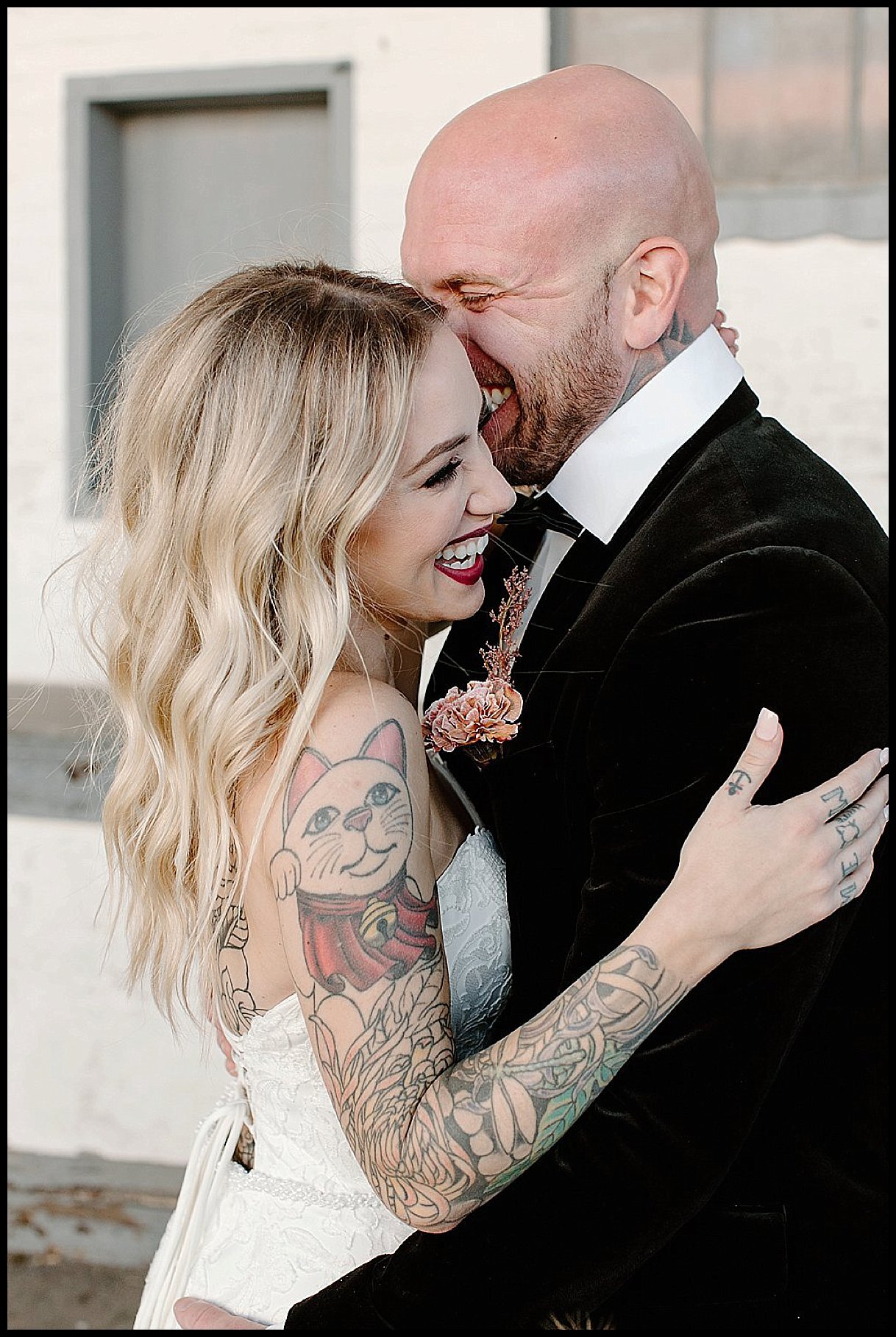 An edgy and alternative bride with tattoos and her groom in a velvet black tuxedo outside Detroit’s The Whiskey Factory. The bride and groom laugh during their wedding photos.