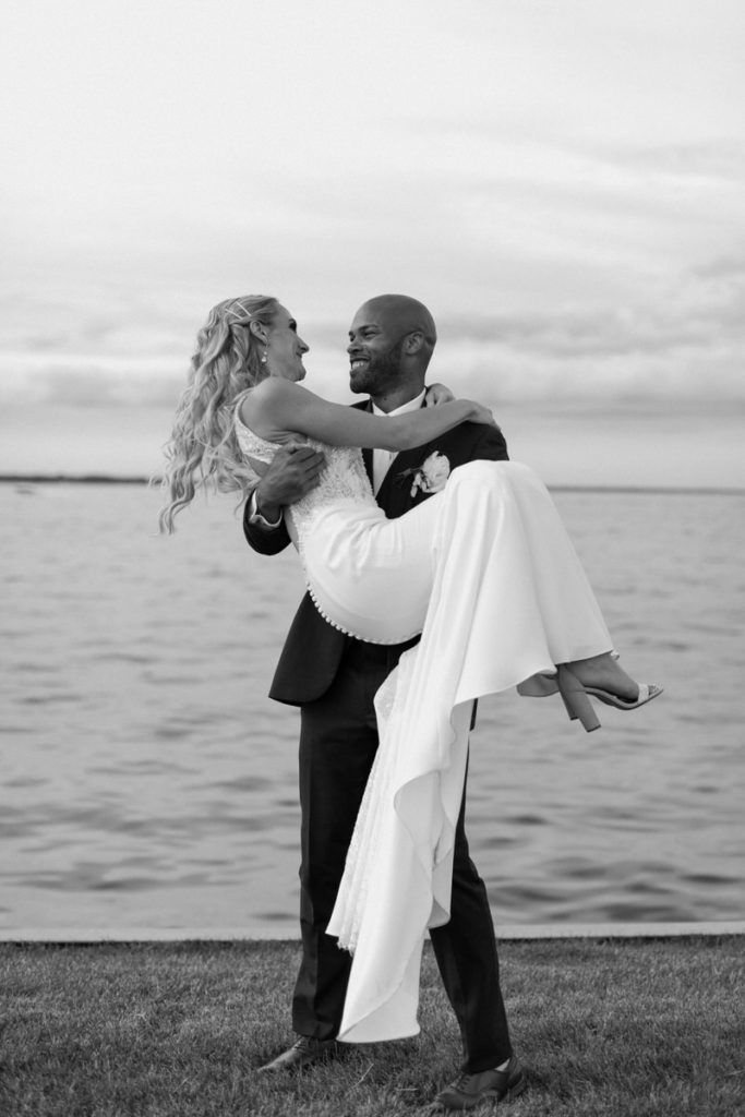 groom holding his bride in his arms on the waters edge