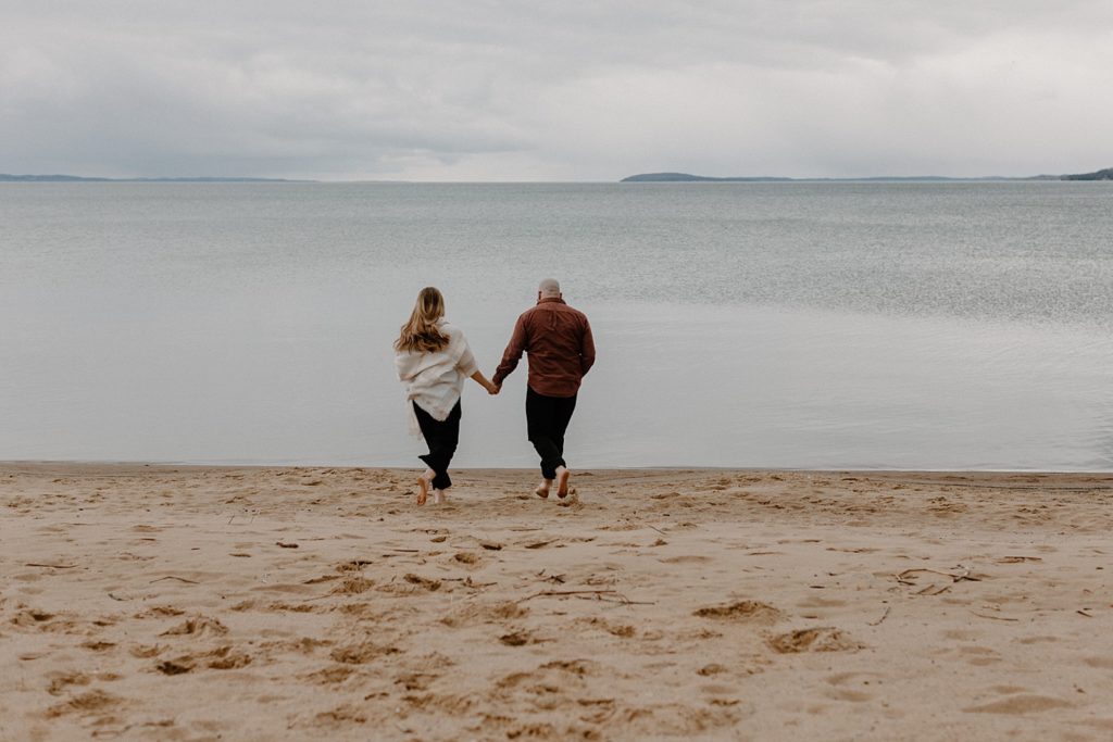 couple holding hands and walking towards the water on a beach 
