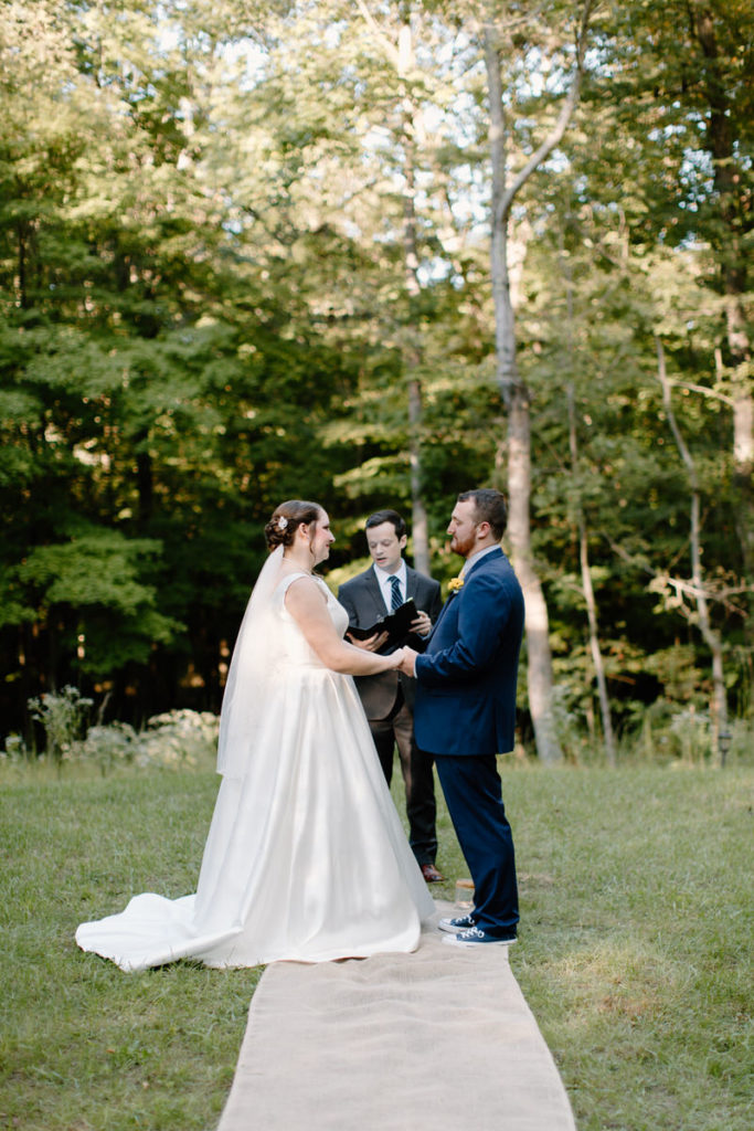 bride and groom holding hands with the officiant behind as they say their vows in a forest