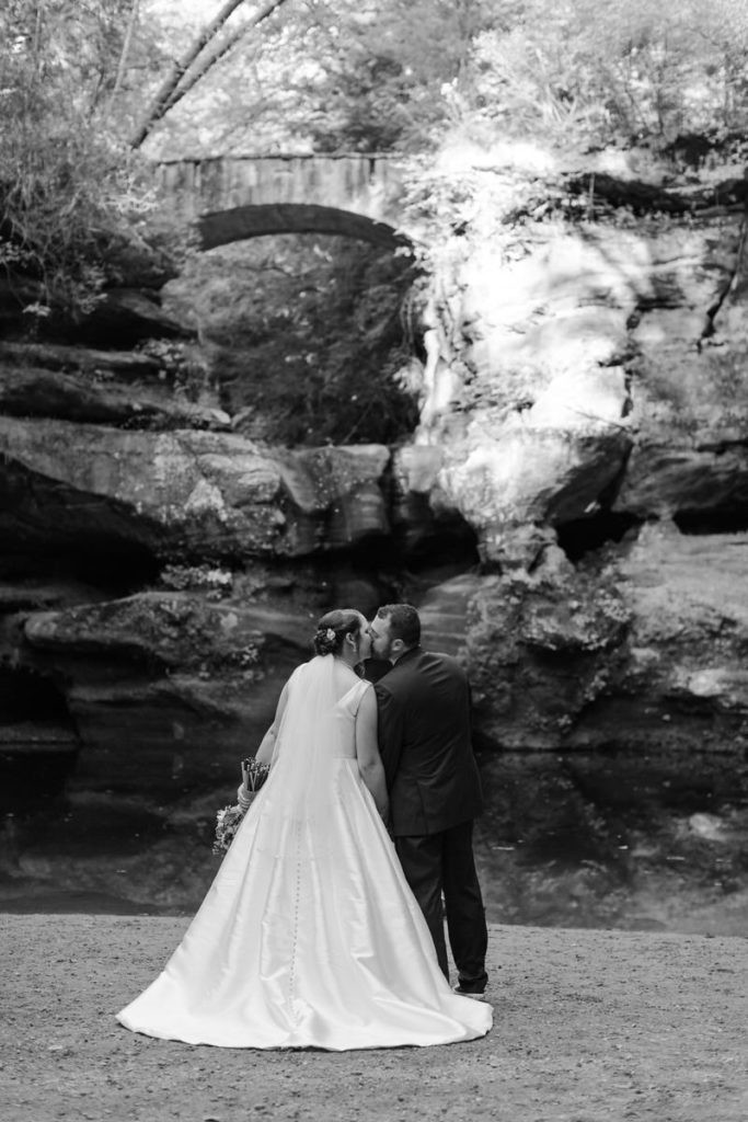 view from behind of bride and groom kissing on water's edge with a large rock wall and bride in the back