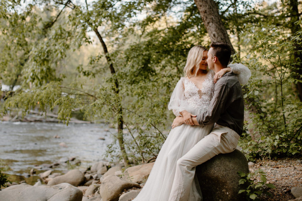 bride sitting on grooms lap on a rocky water's edge