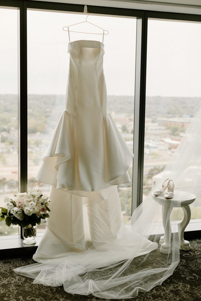 wedding dress handing from large window with shoes on a small table next to it 