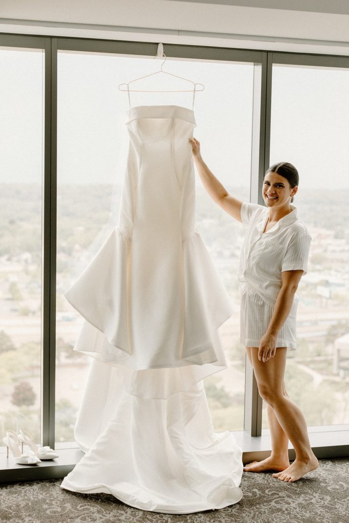 bride smiling while holding wedding dress that is hanging in front of a window 