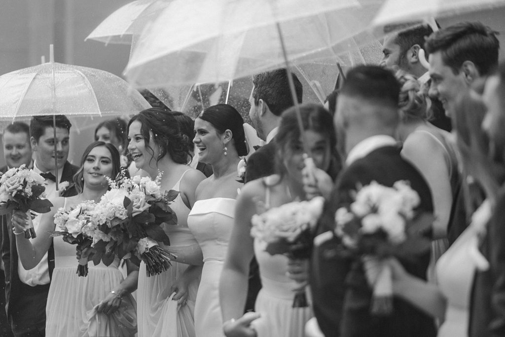 black and white photo of rain falling on wedding party as they stand outside holding umbrellas 
