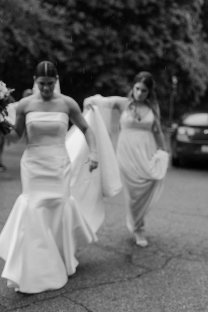 bride walking through the rain while bridesmaids holds up the back of her dress 