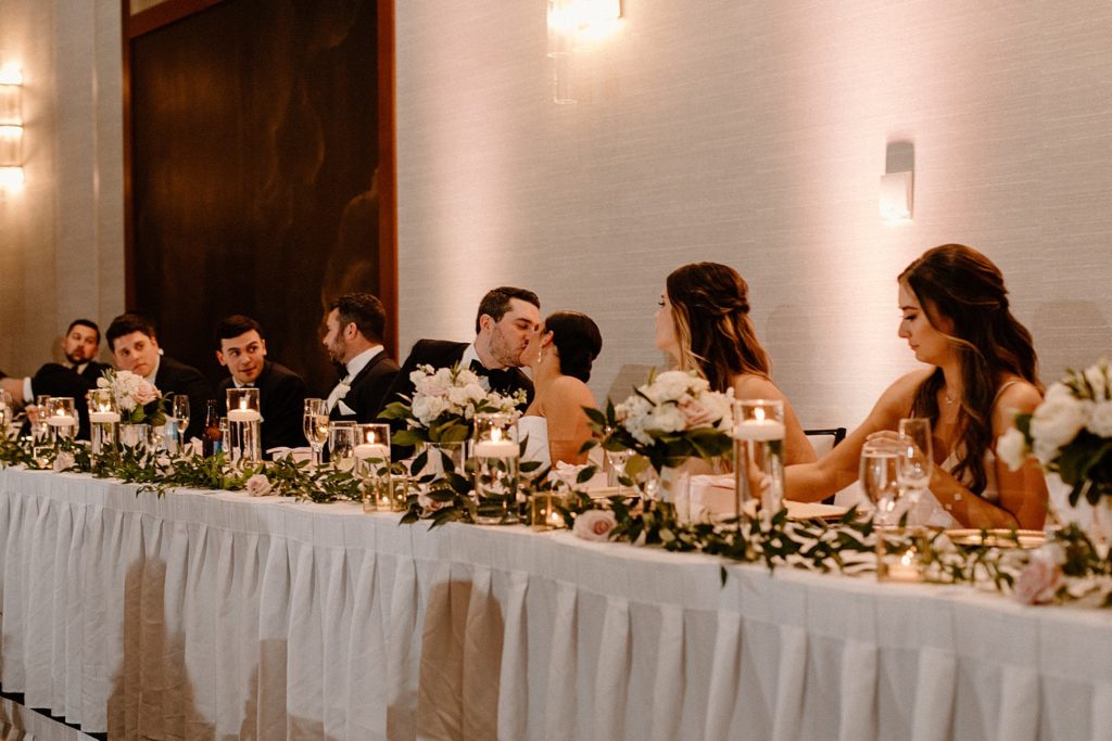 bride and groom kissing while sitting at a reception table with their wedding party 