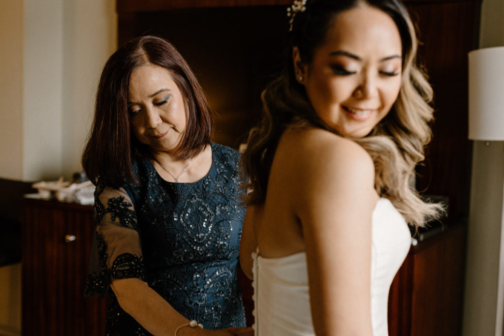 woman helping a bride button up her dress as she looks over her should and smiles 