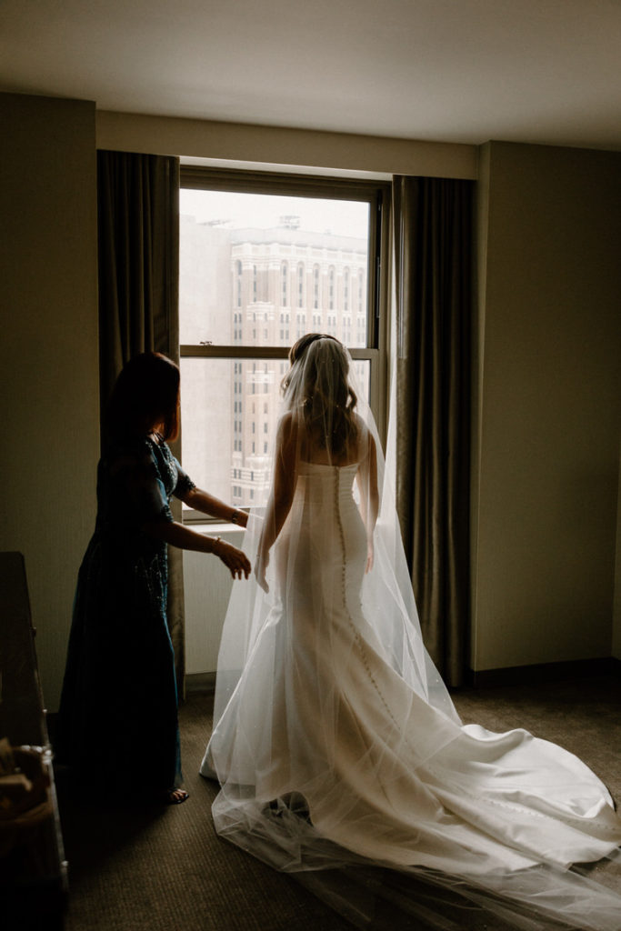 bride standing in front of a window overlooking the city as her mother helps adjust her dress 