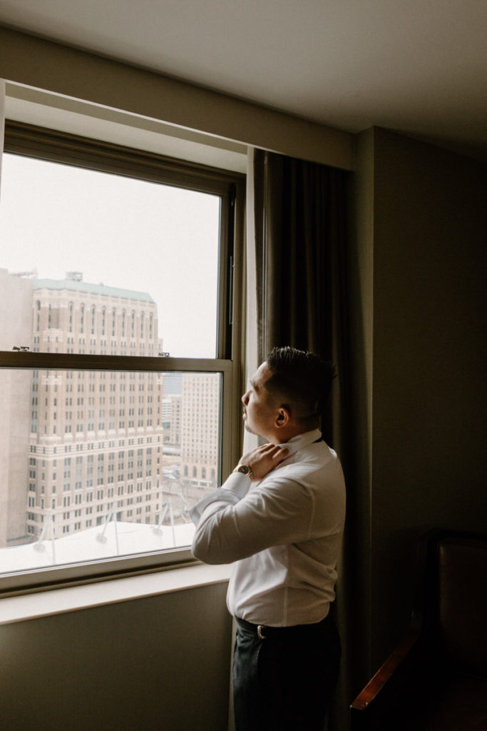 groom looking out of a window that overlooks a city as he adjusts his shirt collar 