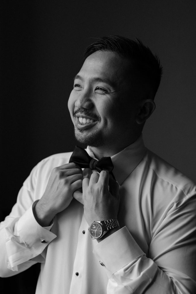 black and white photo of a groom smiling and adjusting his bow tie 