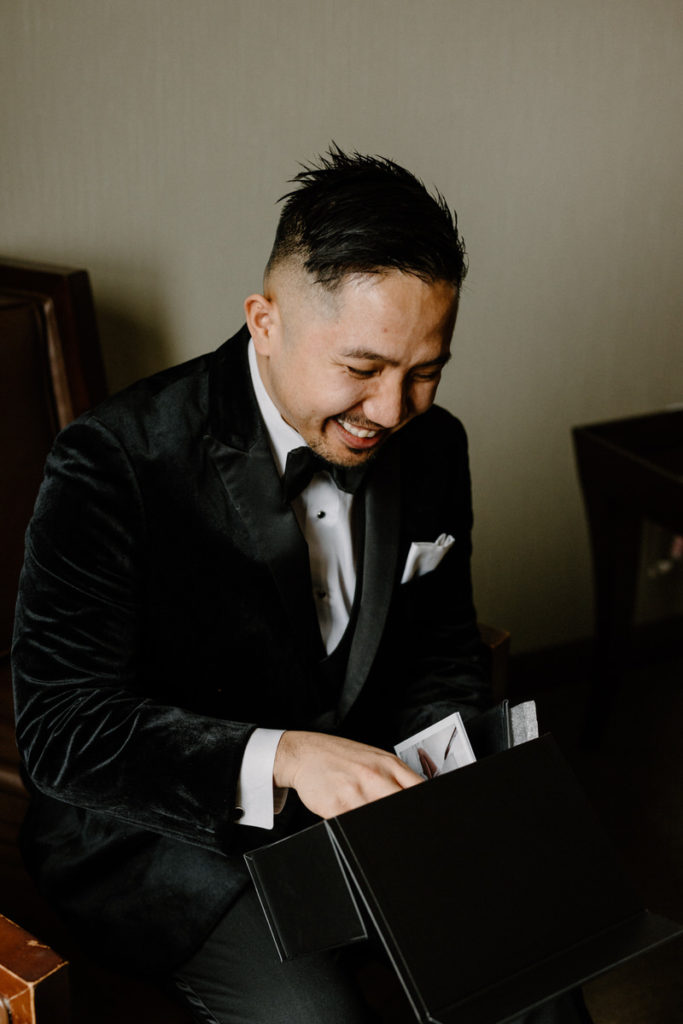 groom sitting down and smiling as he opens a small black box 