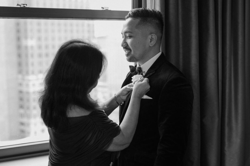 black and white photo of a mom helping pin a boutonniere on her sons wedding suit 