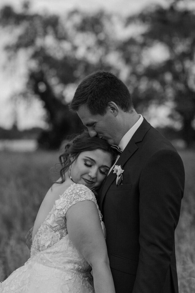 Black and white photo of a bride with her head resting on her grooms chest as they stand outside 