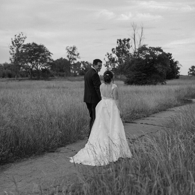 Featured image for is eloping selfish featuring a black and white photo of a couple on their elopement day walking along a path in between two fields