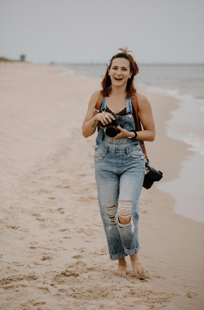 Woman holding a camera with another one hanging from her shoulder as she walks along the edge of the water at a beach. 