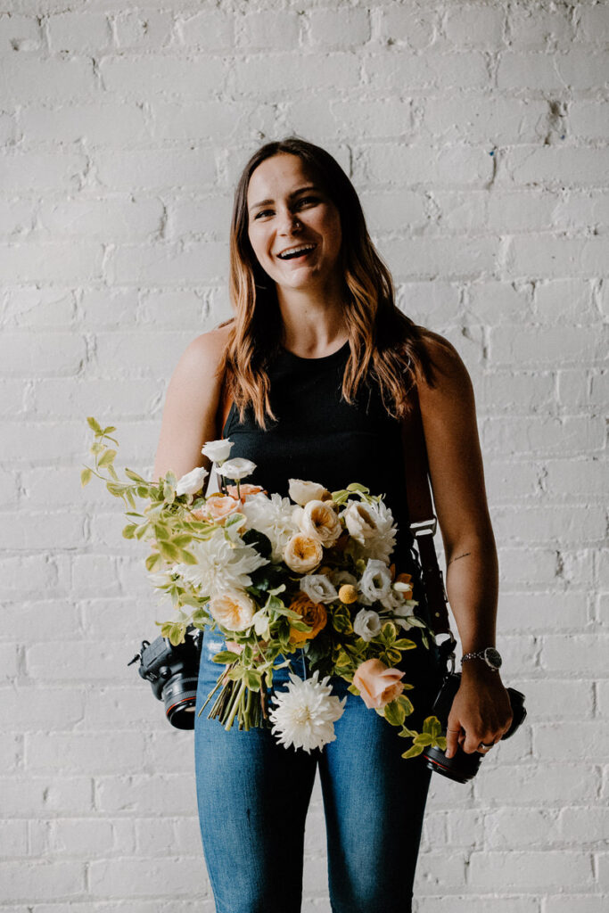 Woman with cameras hanging from both shoulders holding a large bouquet of flowers. 