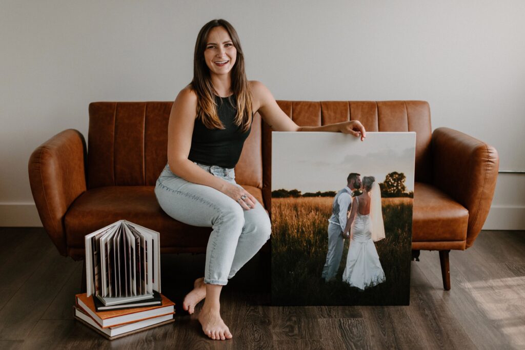 A photographer sitting on a brown leather couch holding up a large canvas photo print of a couple on their wedding day with a stack of wedding albums next to her on the floor. 